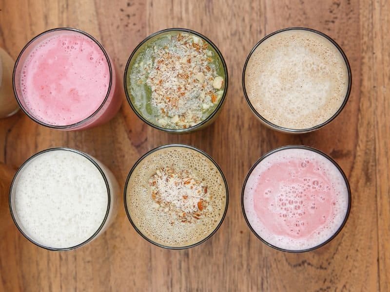 Assorted protein powder shakes in glasses on wooden surface.