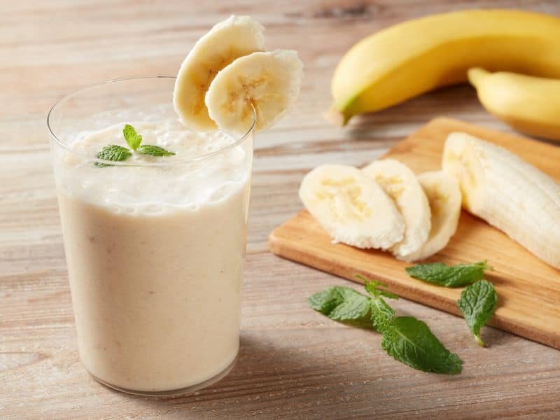 fresh homemade banana protein powder shake in a glass on a kitchen counter with whole bananas and mint leaves