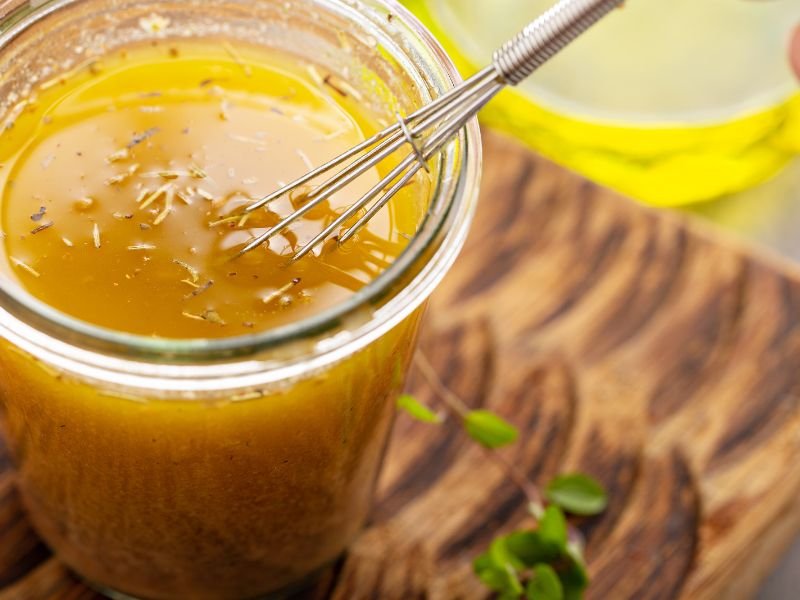 A jar of homemade lemon vinaigrette sits next to whole and cut lemons. In the background, a bowl of fresh greens shows a simple recipe for lemon vinaigrette.