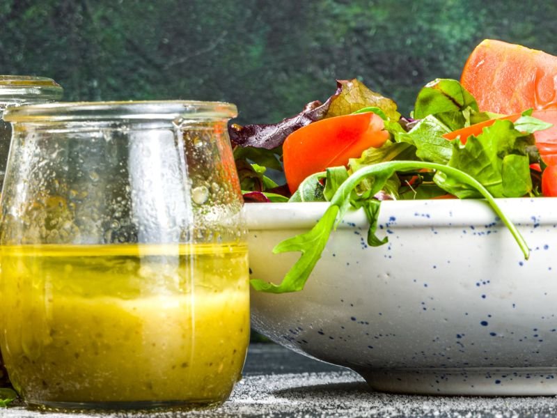 A glass jar of homemade lemon vinaigrette is next to a bowl of fresh salad with greens and sliced tomatoes. This shows a simple lemon vinaigrette recipe.
