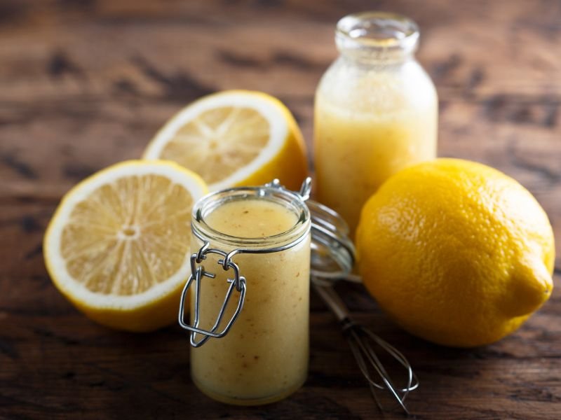 A jar and a bottle of homemade lemon vinaigrette dressing are surrounded by whole and halved lemons on a wooden surface, illustrating an easy lemon vinaigrette recipe.