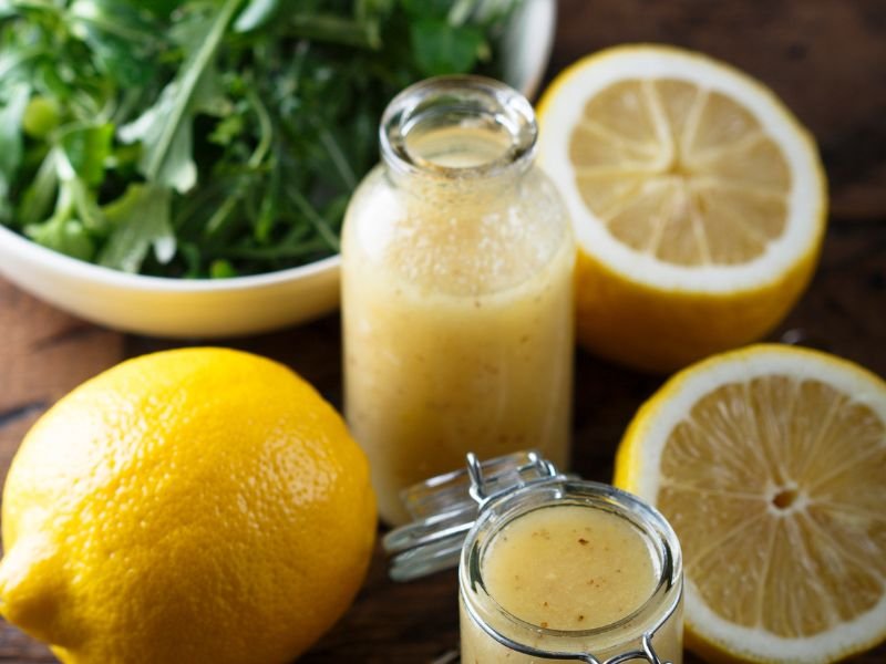 A bottle of homemade lemon vinaigrette stands next to whole and halved lemons, with a bowl of fresh greens behind it. This shows a simple lemon vinaigrette recipe.